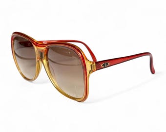 May include: Vintage oversized sunglasses with a red and amber frame and gradient lenses. The sunglasses feature the CD logo on the temples. The lenses transition from a light amber to a darker brown.