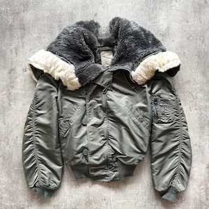 Könnte beinhalten: Eine olivgrüne Bomberjacke mit fellgefütterter Kapuze. Die Jacke hat einen gerippten Saum und Bündchen, mehrere Taschen und Knopfverschlüsse. Die Kapuze hat eine dunkelgraue Fellaußenseite mit cremefarbenem Futter.