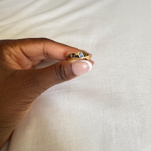 May include: A gold-colored ring with a rectangular blue gemstone and a smaller, dark-colored stone. The ring is held up against a white background.