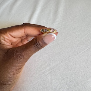 May include: A gold ring with three inset gemstones. The ring has a rectangular shape with a smooth, polished finish. The gemstones are pink and purple, adding a pop of color to the gold band. The ring is held between fingers.