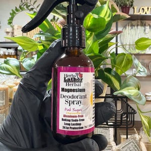 Op de afbeelding: Een bruine glazen fles Herbal Magnesium Deodorant Spray met een zwarte sproeikop. Het etiket leest "Pink Sugar" en "Aluminum-Free". De fles wordt vastgehouden in een zwarte handschoen, met groene planten op de achtergrond.