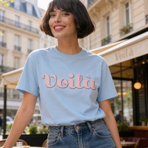 Peut inclure: T-shirt bleu clair avec le mot "Voilà" en rose. Le t-shirt est porté avec un jean bleu. La photo est prise à l'extérieur devant un café.