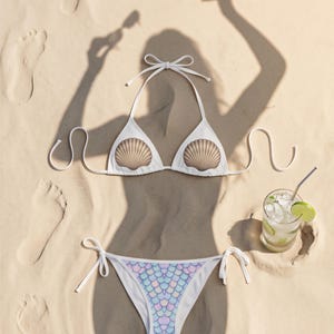 Könnte beinhalten: Ein weißer Bikini mit einem Neckholder-Oberteil, das zwei braune Muschelgrafiken auf den Cups zeigt, und einem Unterteil mit einem pastellfarbenen, irisierenden Meerjungfrauenschuppen-Muster, ausgestattet mit seitlichen Bindebändern.