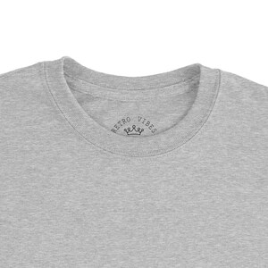 May include: A heather gray crew neck t-shirt. The shirt features the text "RETRO VIBES" with a crown graphic printed on the neckline. The t-shirt is made of a soft, comfortable material.