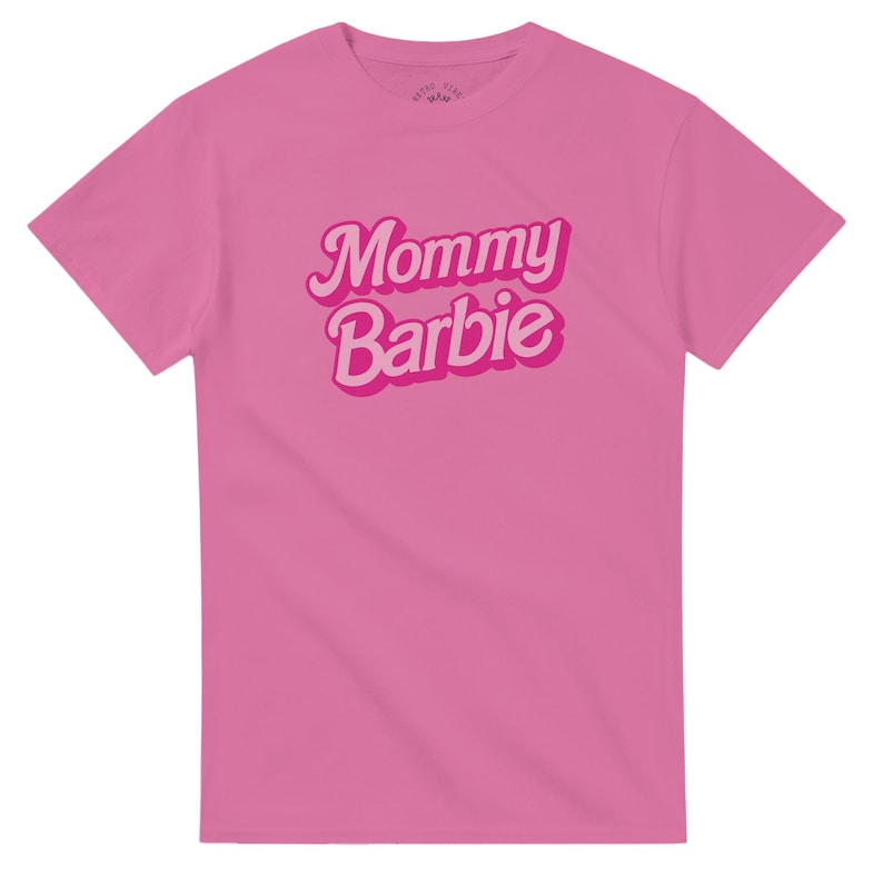May include: A pink t-shirt with the words "Mommy Barbie" in a retro font. The text is in a lighter pink with a darker pink outline. The shirt is a solid pink color.