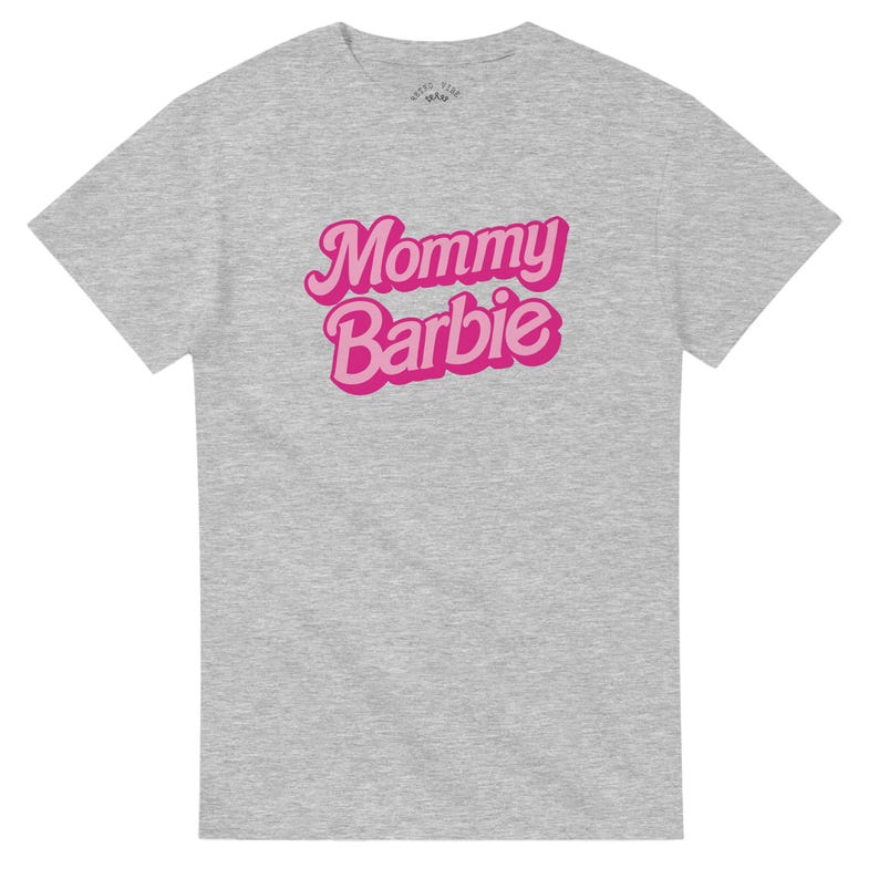 May include: A heather gray t-shirt with the text "Mommy Barbie" in a pink, retro-style font. The shirt has a crew neck and short sleeves. The upper left of the shirt has a small logo.