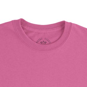 May include: A close-up of a pink t-shirt. The shirt has a crew neck and short sleeves. The text "RETRO VIBES" is printed on the inside collar with a small crown graphic.