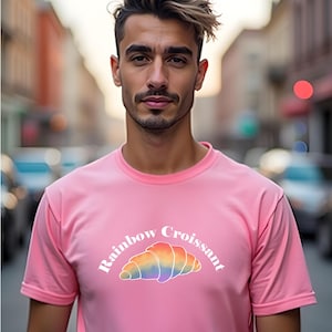 Puede incluir: Una camiseta rosa con la frase "Rainbow Croissant" sobre un gráfico de croissant arcoíris. La camiseta de manga corta está hecha de una tela suave.
