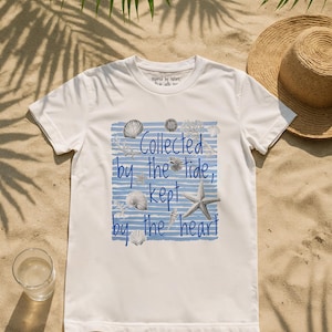 Peut inclure: T-shirt blanc avec un motif sur le thème de la plage. Le t-shirt présente des rayures horizontales bleues, des coquillages, une étoile de mer et le texte "Collected by the tide, kept by the heart". Le t-shirt est sur une surface sablonneuse.