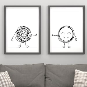 May include: Two framed art prints. One print features a black scribble figure with arms and legs. The other print shows a smiling circle figure with arms and legs. Both prints are in gray frames and hang above a gray sofa.