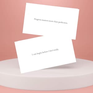 May include: Two white cards with inspirational quotes on a white pedestal against a light pink background. The top card reads "Progress matters more than perfection." The bottom card reads "I can begin before I feel ready."