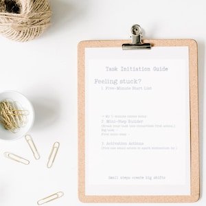 May include: A corkboard clipboard with a white sheet of paper attached. The paper has the text "Task Initiation Guide" and "Feeling stuck?" in a light purple font. The paper also has a list of steps with the headings "Five-Minute Start List", "Mini-Step Builder", and "Activation Actions". The text at the bottom of the paper reads "Small steps create big shifts".