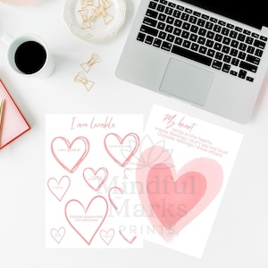 May include: A workspace scene featuring two printable affirmation sheets with heart designs and text. The words "I am lovable" and "My heart" are visible. A laptop, coffee cup, and pink notebook are also on the white surface.