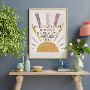 Pode incluir: Impressão artística emoldurada com o texto "YOU ARE ALLOWED TO IMAGINE THE BEST CASE SCENARIO TOO". A obra apresenta um desenho de sol em tons de castanho, bege e amarelo. A impressão é exibida numa prateleira de madeira com vasos e plantas decorativas.