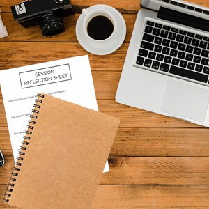 Peut inclure: Une feuille de papier blanche avec le texte "SESSION REFLECTION SHEET" sur une table en bois. Il y a un carnet brun avec une reliure spirale, un stylo, un ordinateur portable, une tasse à café et un appareil photo sur la table.