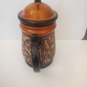 Peut inclure: Une chope à bière en bois avec couvercle et anse. La chope est marron avec des motifs floraux sculptés et des accents noirs. Le couvercle est surmonté d'un fleuron noir.