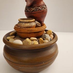 Peut inclure: Une fontaine d'intérieur décorative avec un design à plusieurs niveaux. La fontaine comprend une grande base en céramique marron, un niveau intermédiaire avec un petit pot et un niveau supérieur avec une cruche marron. La fontaine est ornée de petites pierres lisses.