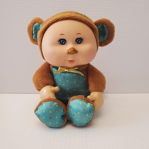 Peut inclure: Une poupée vintage habillée en singe, avec un chapeau marron et des détails d'oreilles bleus. La poupée a les yeux bleus et une expression souriante. Elle porte une combinaison turquoise à pois jaunes et un nœud jaune.