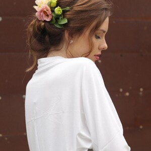May include: A woman with brown hair styled up with a floral hairpiece. She is wearing a white top with a subtle bird design on the back. The background is a dark brown, creating a contrast with the white clothing.
