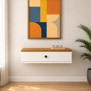 Puede incluir: Una consola flotante montada en la pared con cuerpo blanco y tapa de madera natural, que presenta un único pomo circular negro. Sobre la consola cuelga una pieza de arte mural rectangular enmarcada con formas geométricas en azul, naranja y amarillo.