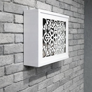 May include: A white wall-mounted cabinet with a decorative, openwork design on the front. The intricate pattern is cut into the white surface, revealing a darker interior. The cabinet is mounted on a gray brick wall.