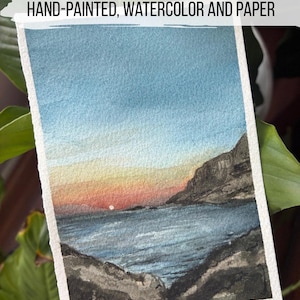 May include: Hand-painted watercolor landscape art on paper. The artwork depicts a sunset over a body of water, with mountains on either side. The sky transitions from blue to orange and red. The paper has a white border.
