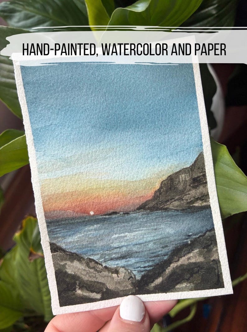 May include: Hand-painted watercolor landscape art on paper. The artwork depicts a sunset over a body of water, with mountains on either side. The sky transitions from blue to orange and red. The paper has a white border.