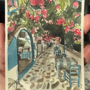 May include: Watercolor painting of a European street scene. A cobblestone path leads through a shaded area with tables and chairs. Red flowers and green foliage hang overhead. Buildings are painted in white and blue.