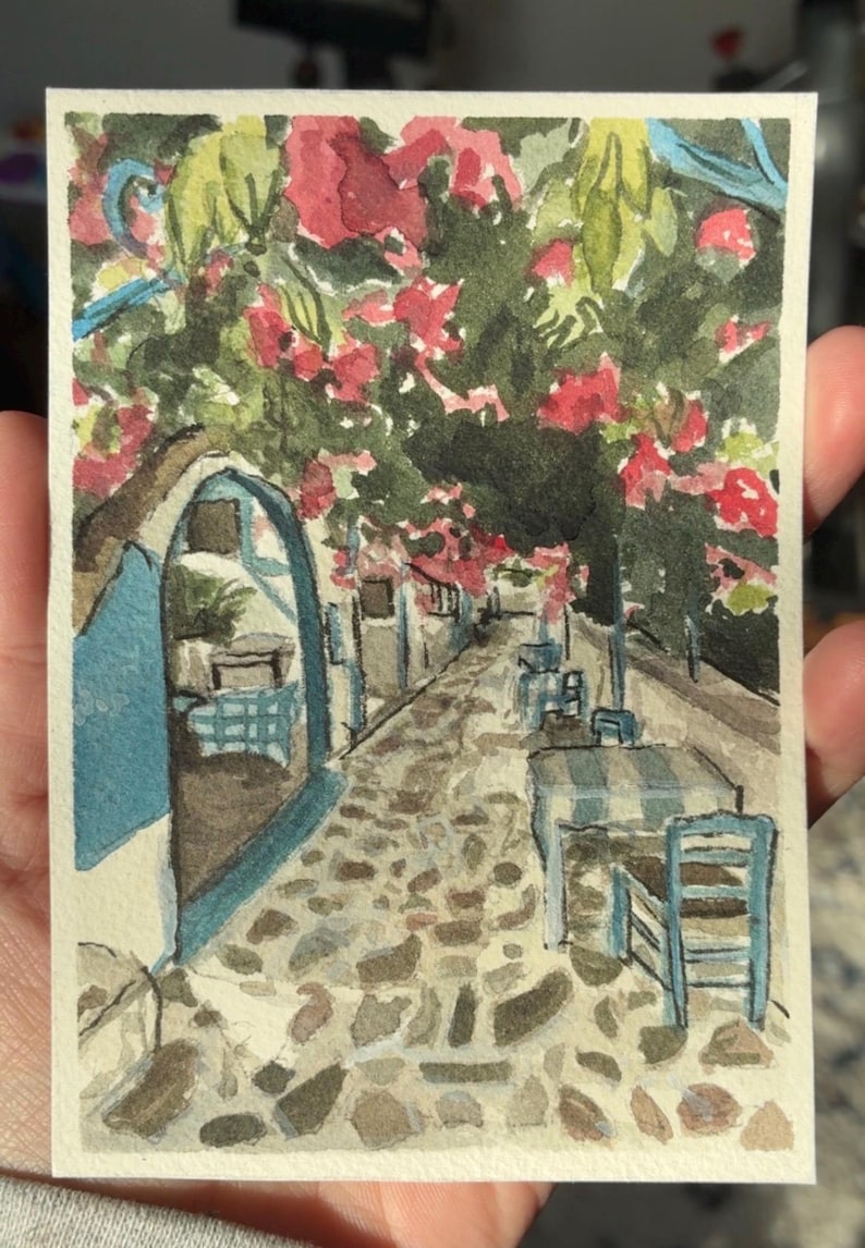 May include: Watercolor painting of a European street scene. A cobblestone path leads through a shaded area with tables and chairs. Red flowers and green foliage hang overhead. Buildings are painted in white and blue.
