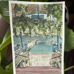 May include: Watercolor painting of a lakeside scene with a dock, water, and lush greenery. The artwork features a blue and brown building, a stone walkway, and a serene atmosphere. The painting is on a rectangular piece of paper.