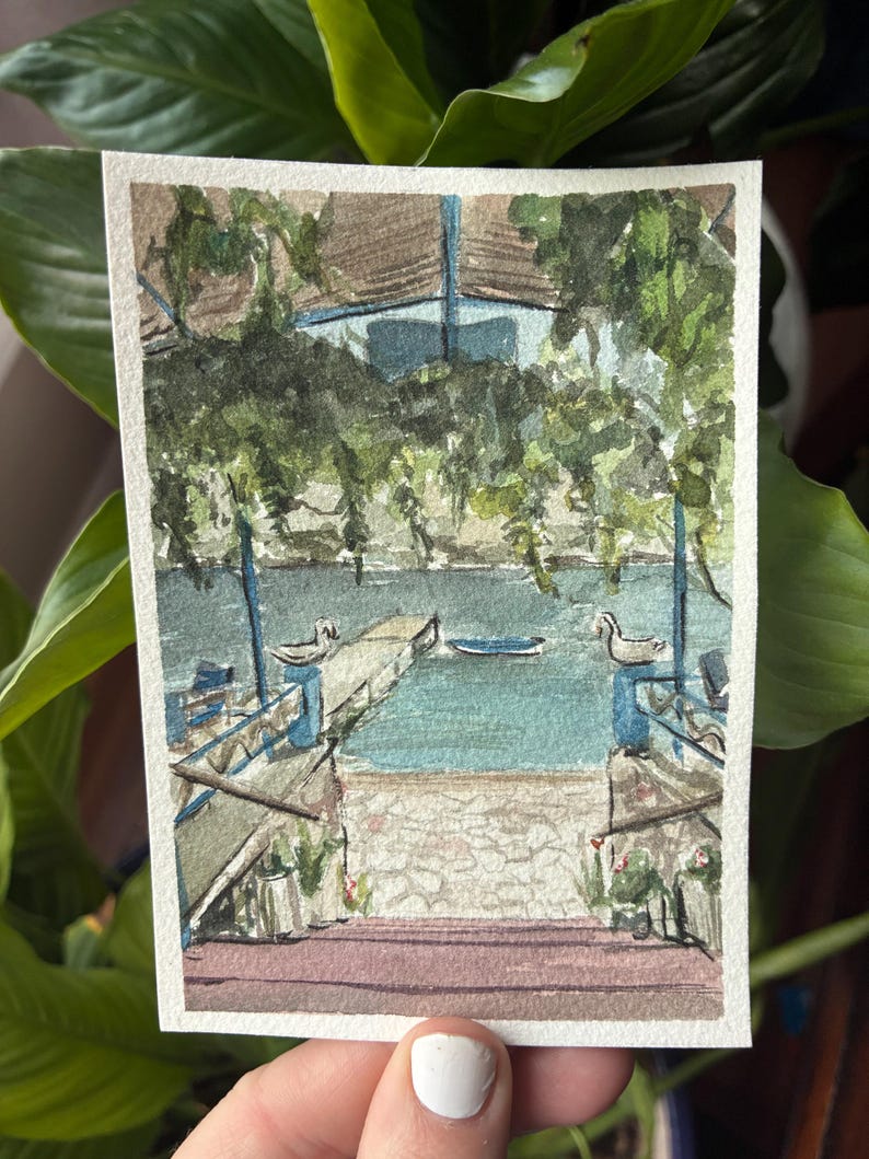 May include: Watercolor painting of a lakeside scene with a dock, water, and lush greenery. The artwork features a blue and brown building, a stone walkway, and a serene atmosphere. The painting is on a rectangular piece of paper.