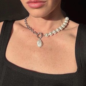 Puede incluir: Un collar plateado con una combinación de eslabones de cadena y perlas blancas grandes y redondas. Un colgante de perla grande e irregular cuelga de un cierre circular. El collar se usa sobre una camiseta negra.