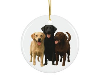 Könnte beinhalten: Runder, weißer Keramikschmuck mit drei Labrador Retrievern: einem gelben, einem schwarzen und einem schokoladenbraunen. Die Hunde haben die Zungen draußen. Ein goldener Faden zum Aufhängen ist befestigt. Ein dekoratives Ornament mit Haustier-Motiv.