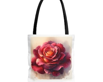 Op de afbeelding: Een witte tote bag met zwarte handvatten heeft een aquarel schilderij van een enkele rode roos met gele bloemblaadjes in het midden. De roos is afgebeeld met waterdruppels op de bloemblaadjes.