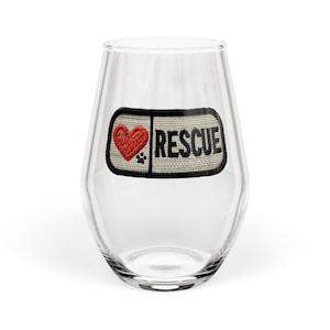 May include: A clear stemless wine glass with a rectangular patch featuring a red heart, paw print, and the word "RESCUE" in black. The patch is outlined in black and has a beige background.