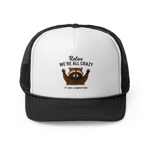 Puede incluir: Gorra de camionero en blanco y negro con un diseño de mapache. El mapache tiene los brazos levantados y el texto dice "Relax we're all crazy, it's not a competition".