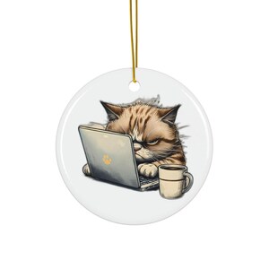 May include: A round, white ceramic ornament depicting a grumpy cat using a laptop. The cat has brown and tan fur and is next to a coffee mug. A gold string is attached for hanging.