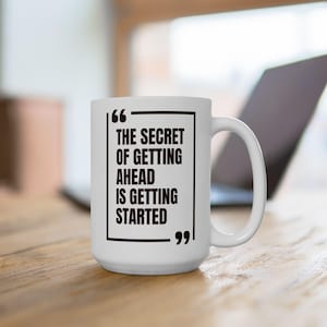 May include: White ceramic mug with a black rectangular frame containing the quote: "THE SECRET OF GETTING AHEAD IS GETTING STARTED." The mug has a large handle and is set on a wooden surface, with a laptop in the background.