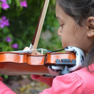 Puede incluir: Una persona joven tocando un viol&iacute;n marr&oacute;n con un reposabrazos blanco y negro. El viol&iacute;n tiene un cuello de madera y un reposabrazos negro.
