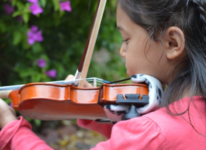 Puede incluir: Una persona joven tocando un viol&iacute;n marr&oacute;n con un reposabrazos blanco y negro. El viol&iacute;n tiene un cuello de madera y un reposabrazos negro.