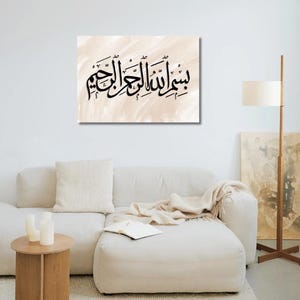 May include: A canvas print featuring black Arabic calligraphy on a beige background. The artwork is displayed above a white sectional sofa in a living room setting. The room includes a wooden side table, a floor lamp, and a neutral-toned rug.