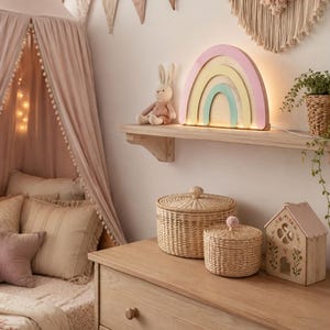 Peut inclure: Une lampe en bois en forme d'arc-en-ciel avec des arches pastel rose, jaune et menthe est posée sur une étagère murale à côté d'un lapin en peluche. Deux paniers ronds tressés avec couvercles et une boîte en bois en forme de maison.