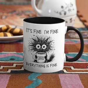 以下が含まれることがあります： 黒い内側とハンドルが付いた白いコーヒーマグ。マグには、コーヒーカップを持った黒い毛むくじゃらの生き物の漫画イラストが描かれており、テキストは「IT'S FINE I'M FINE EVERYTHING IS FINE」です。