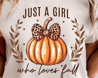 May include: Cream-colored t-shirt featuring a large orange pumpkin with a leopard print bow. The text "JUST A GIRL who loves fall" is printed above and below the pumpkin, surrounded by brown leaves and stars.