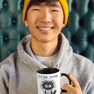 以下が含まれることがあります： 黒い内側とハンドルが付いた白いコーヒーマグ。マグには「IT'S FINE I'M FINE EVERYTHING IS FINE」の文字と、コーヒーカップを持った漫画のモンスターが描かれています。マグは、グレーのパーカーと黄色のビーニーをかぶった人が持っています。
