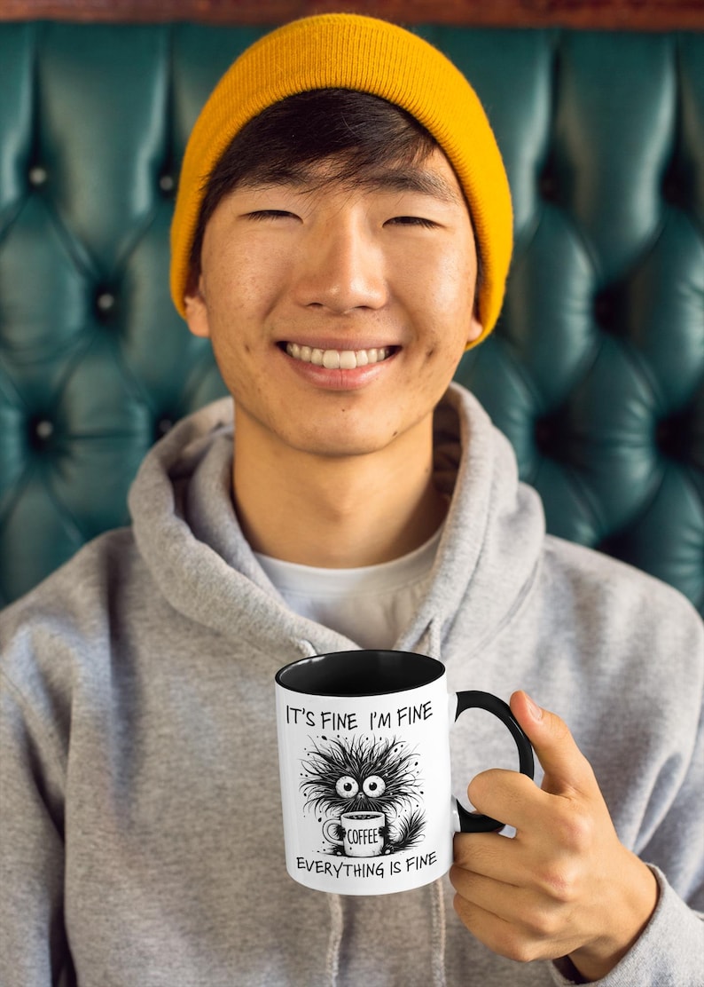 以下が含まれることがあります： 黒い内側とハンドルが付いた白いコーヒーマグ。マグには「IT'S FINE I'M FINE EVERYTHING IS FINE」の文字と、コーヒーカップを持った漫画のモンスターが描かれています。マグは、グレーのパーカーと黄色のビーニーをかぶった人が持っています。