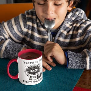 以下が含まれることがあります： 赤い内側とハンドルが付いた白いコーヒーマグ。コーヒーカップを持った漫画のモンスターが描かれています。マグカップのテキストは「IT'S FINE I'M FINE EVERYTHING IS FINE」です。マグカップは青い表面にあります。