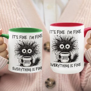 以下が含まれることがあります： 白地のコーヒーマグ2つ。それぞれ、大きな目とコーヒーカップを持つ、黒くて毛むくじゃらの生き物が描かれた漫画のデザインです。「IT'S FINE I'M FINE EVERYTHING IS FINE」の文字が各マグにプリントされています。