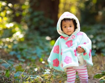 Könnte beinhalten: Ein hellblauer Kapuzenumhang mit einem wiederkehrenden Muster aus rosa Blüten mit weißen Zentren. Der Umhang hat ein weißes Fleecefutter und einen einzelnen Knopfverschluss am Hals. Er wird über einer weißen Leggings mit horizontalen rosa Streifen getragen.