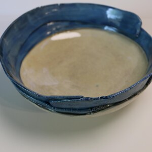 May include: A handmade ceramic bowl with a wide, irregular rim. The bowl features a light beige interior and a contrasting dark blue exterior rim. The rim has a textured, uneven surface, adding to its unique, artistic appeal.